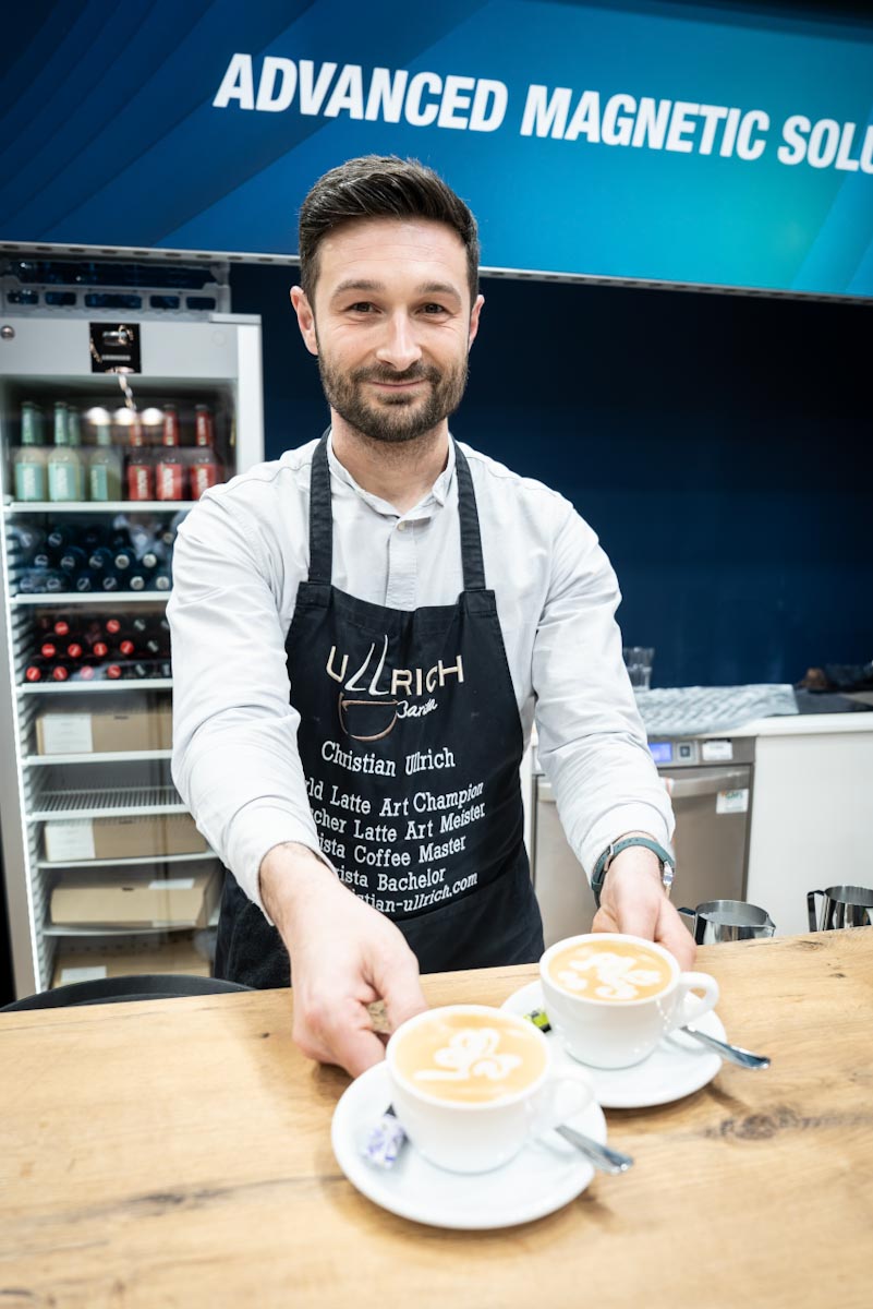 barista-catering-cappuccino-latte-art-messestand.jpg Christian Ullrich reicht zwei Cappuccino mit Latte Art über die Kaffeebar am Messestand